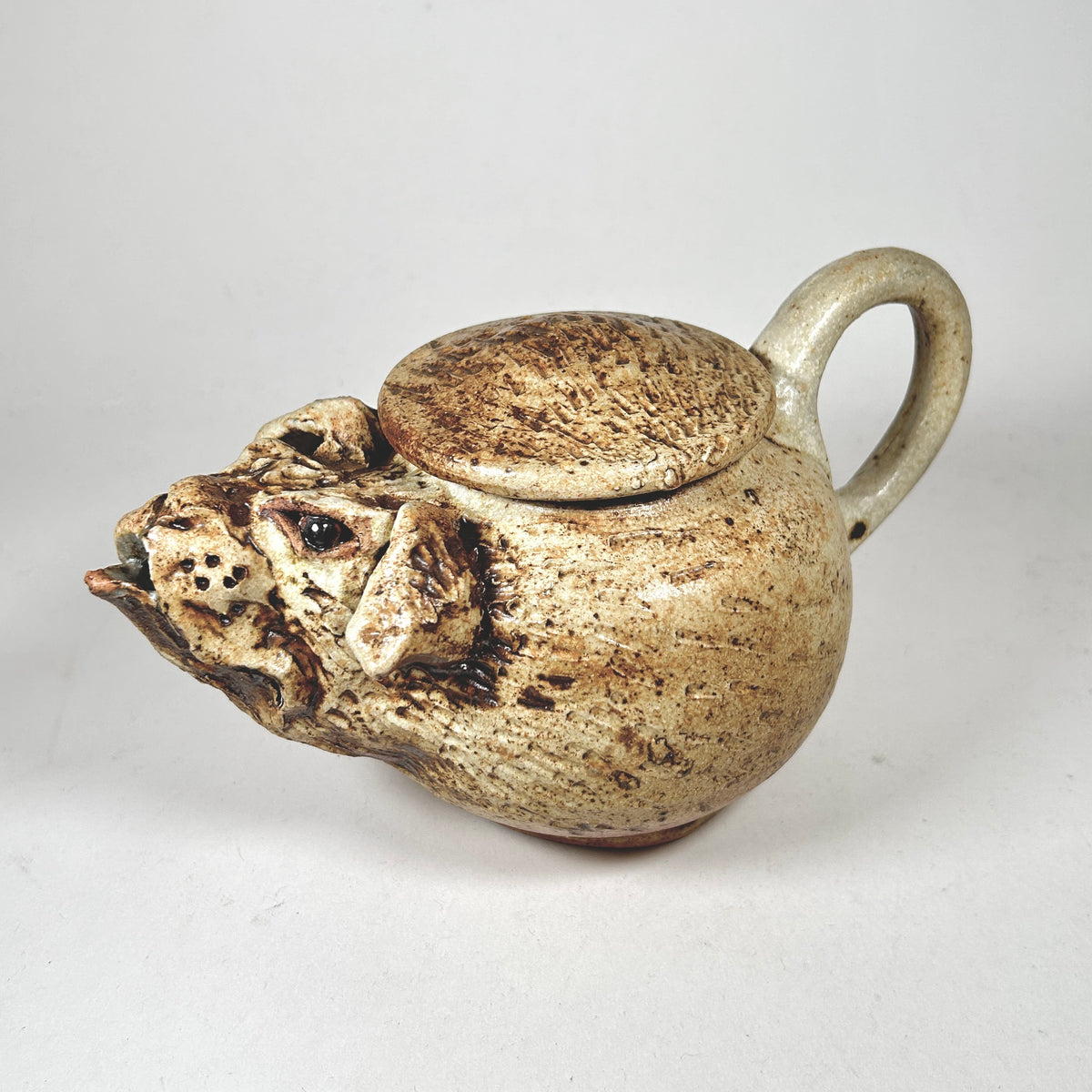 A handmade ceramic teapot with a sculpted animal head spout and a matching lid, featuring a textured beige and brown glaze on a neutral light gray background.